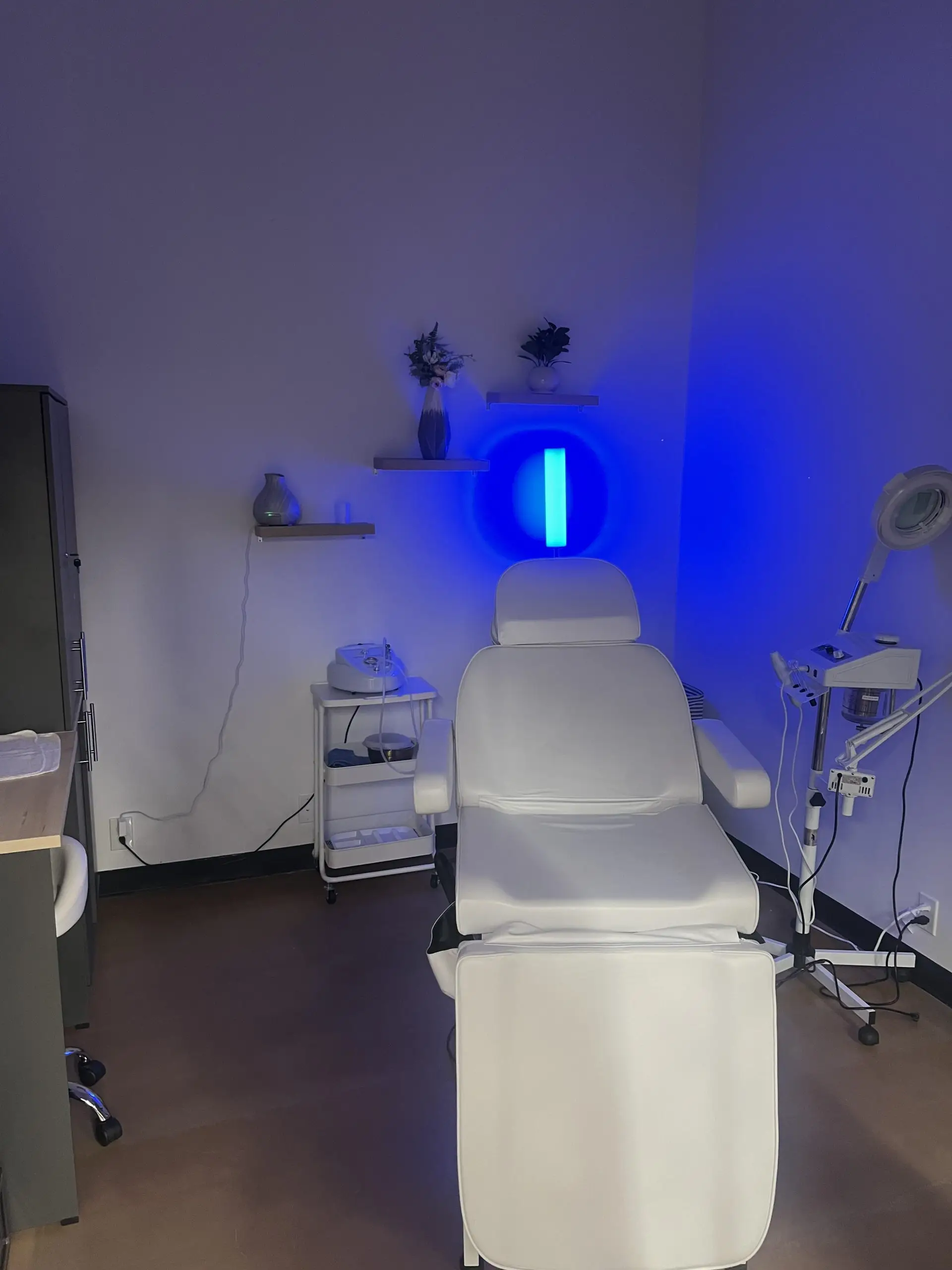 Private esthetic treatment suite featuring a white treatment bed, blue ambient lighting, and various skincare equipment.