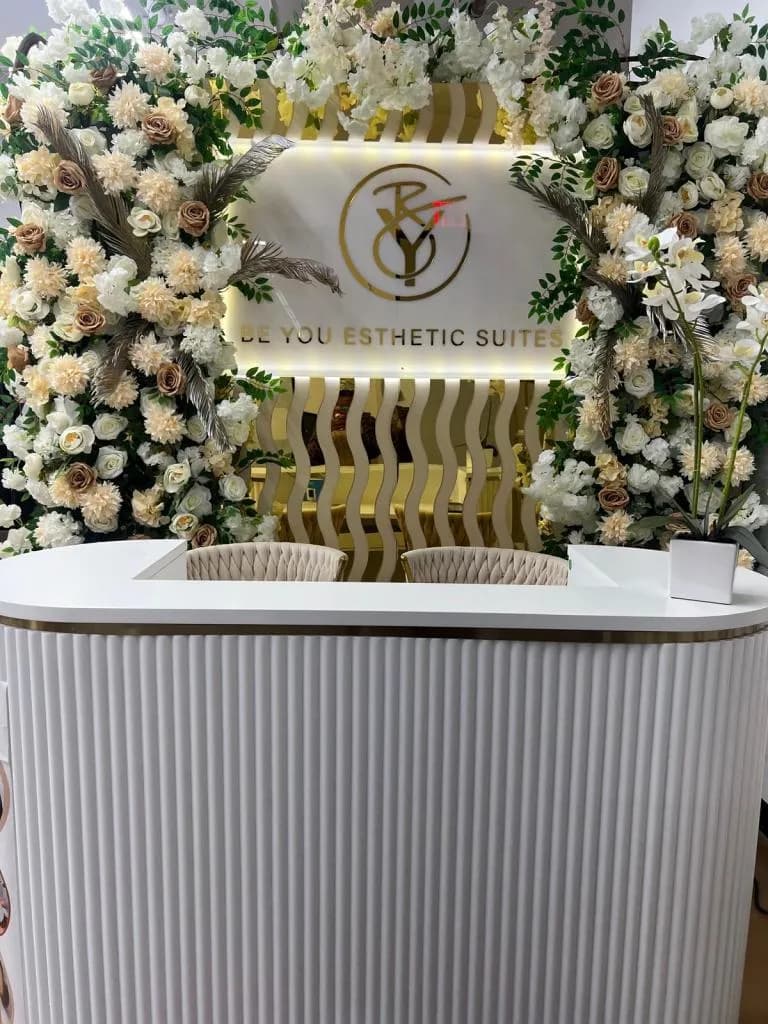 Elegant reception area at Be You Esthetic Suites, featuring a floral backdrop, modern design elements, and the brand logo.